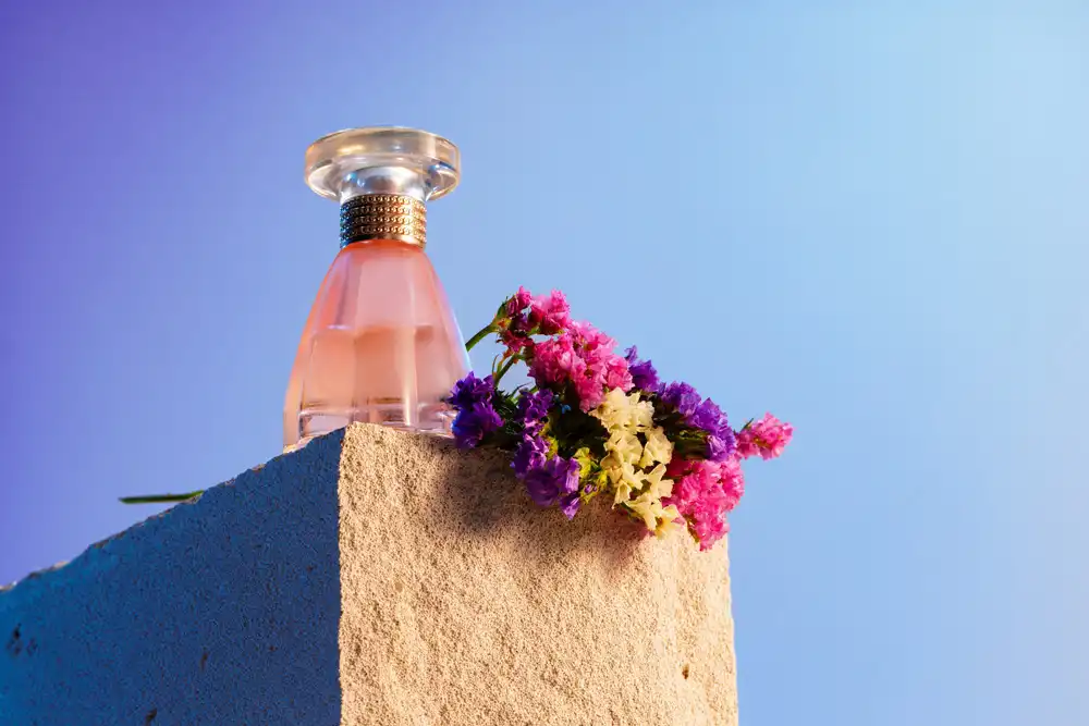 شیشه عطر با سر گرد روی سنگ در کنار گل‌ها رنگی